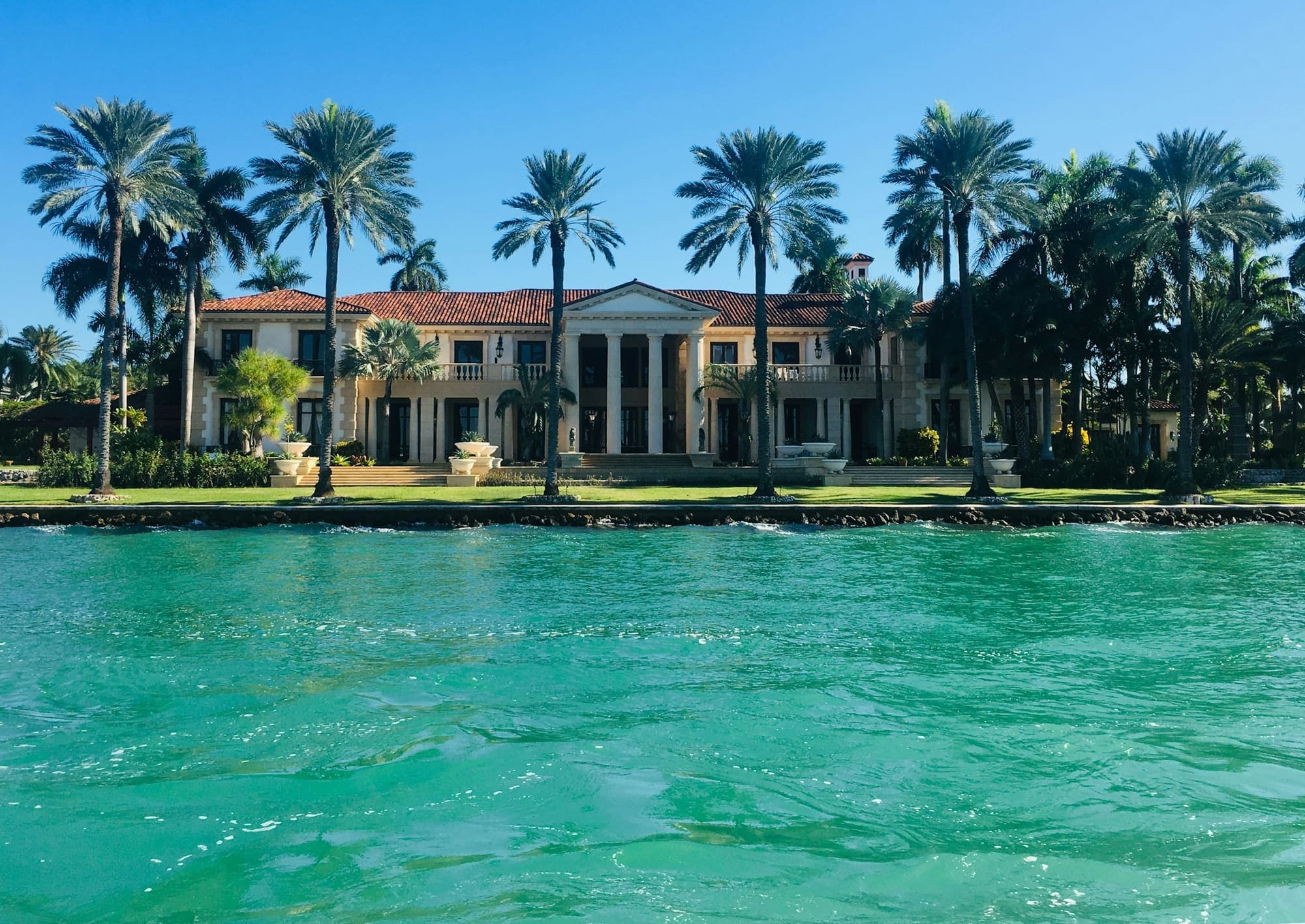 Waterfront estate behind a row of royal palms across turquoise water